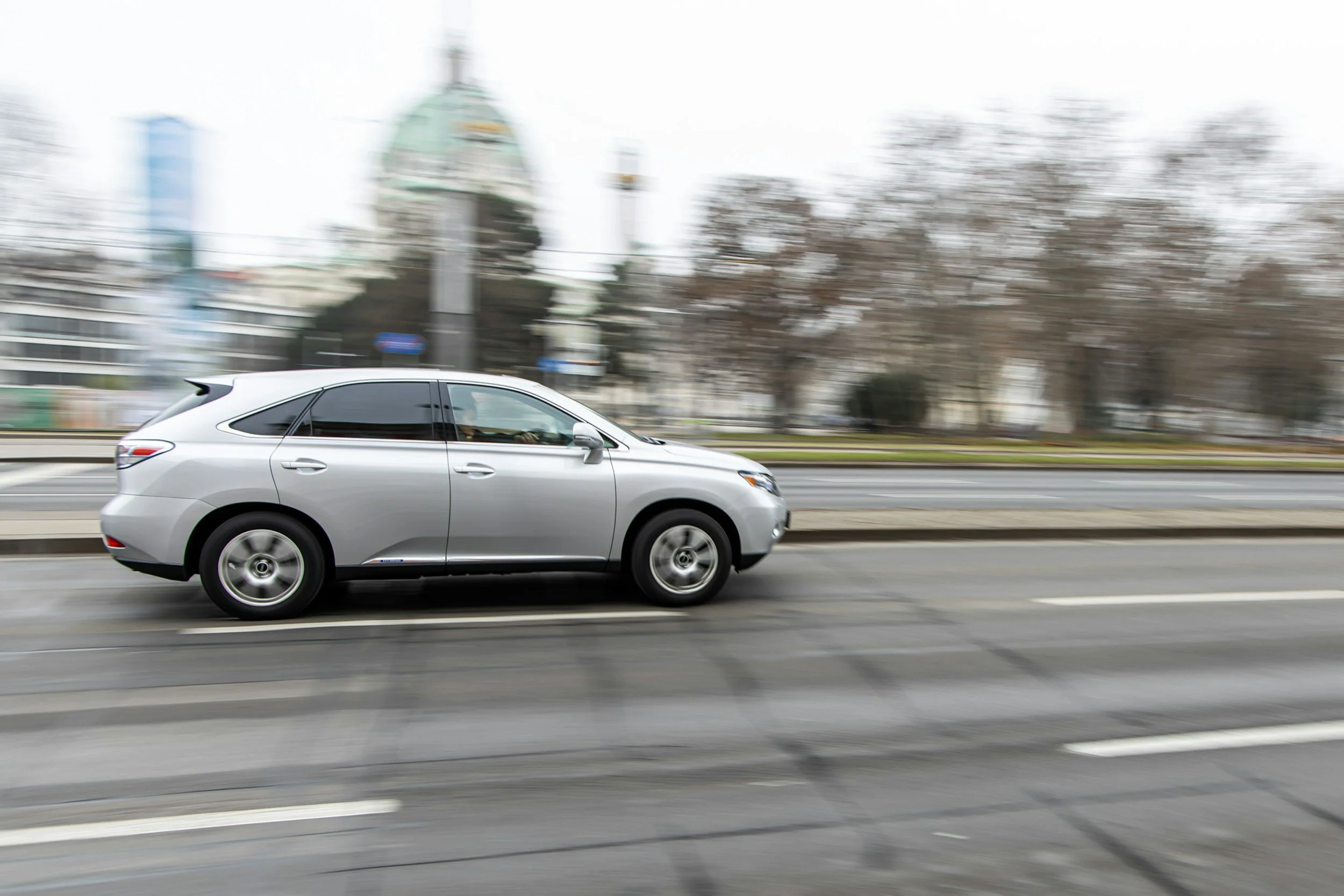 A silver car driving on a city street with blurred background of buildings and trees.