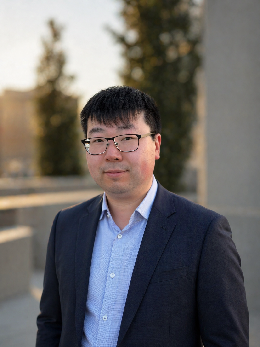 A portrait of a man wearing glasses, a dark blazer, and a light blue button-up shirt, standing outdoors with trees and a building in the background.