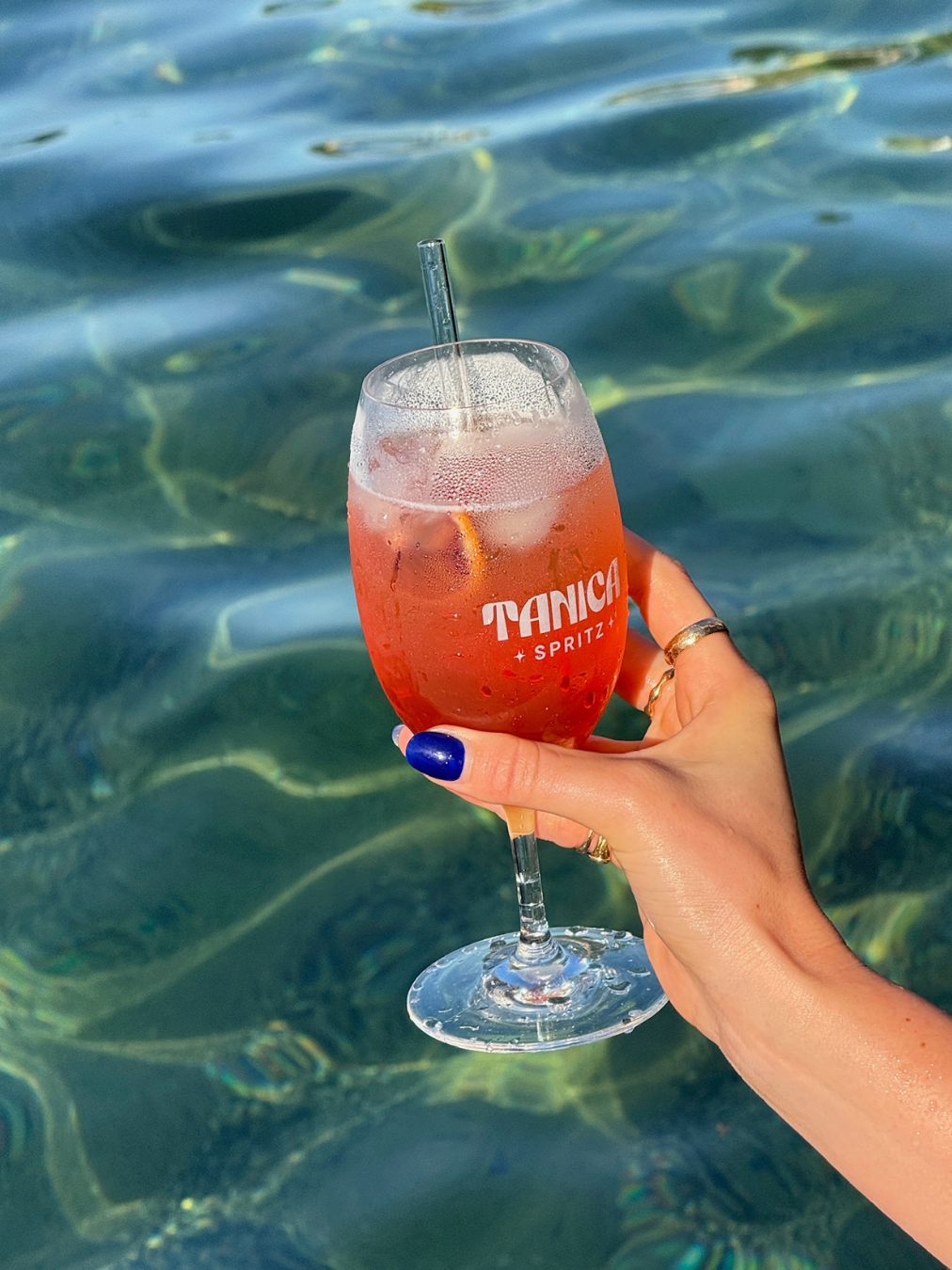 A hand with blue nails and gold rings holding a glass of pink drink with ice and a lemon slice, labeled 'Tanica Spritz,' against a body of water.