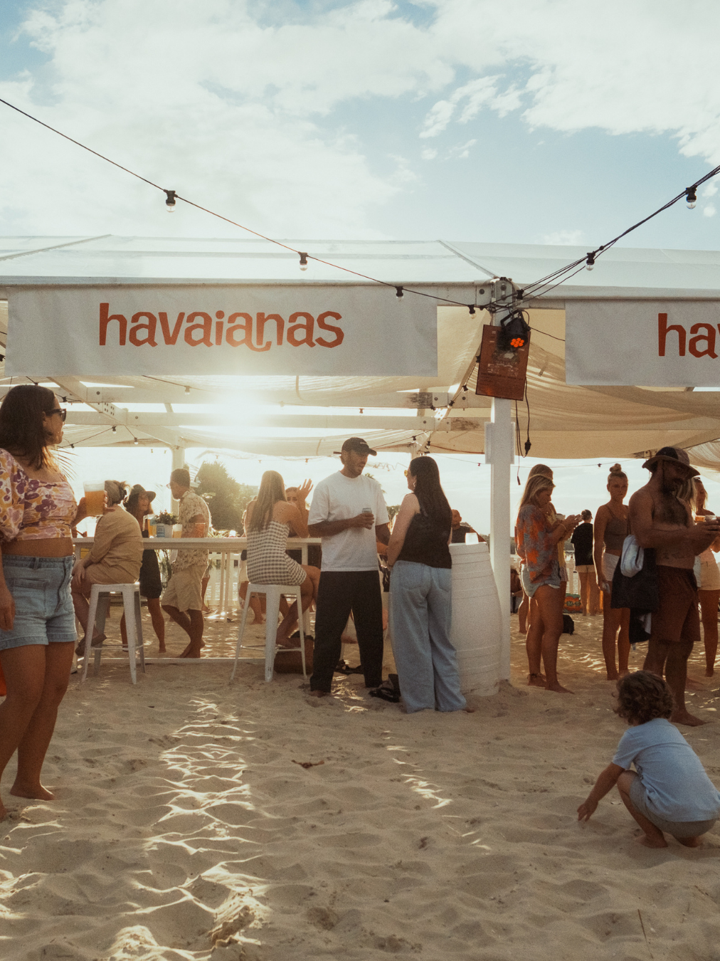 A beach scene with a Haleiwa's food stand, people socializing, some sitting on barstools, and others standing or crouching on the sand, with bright sunlight and a partly cloudy sky.