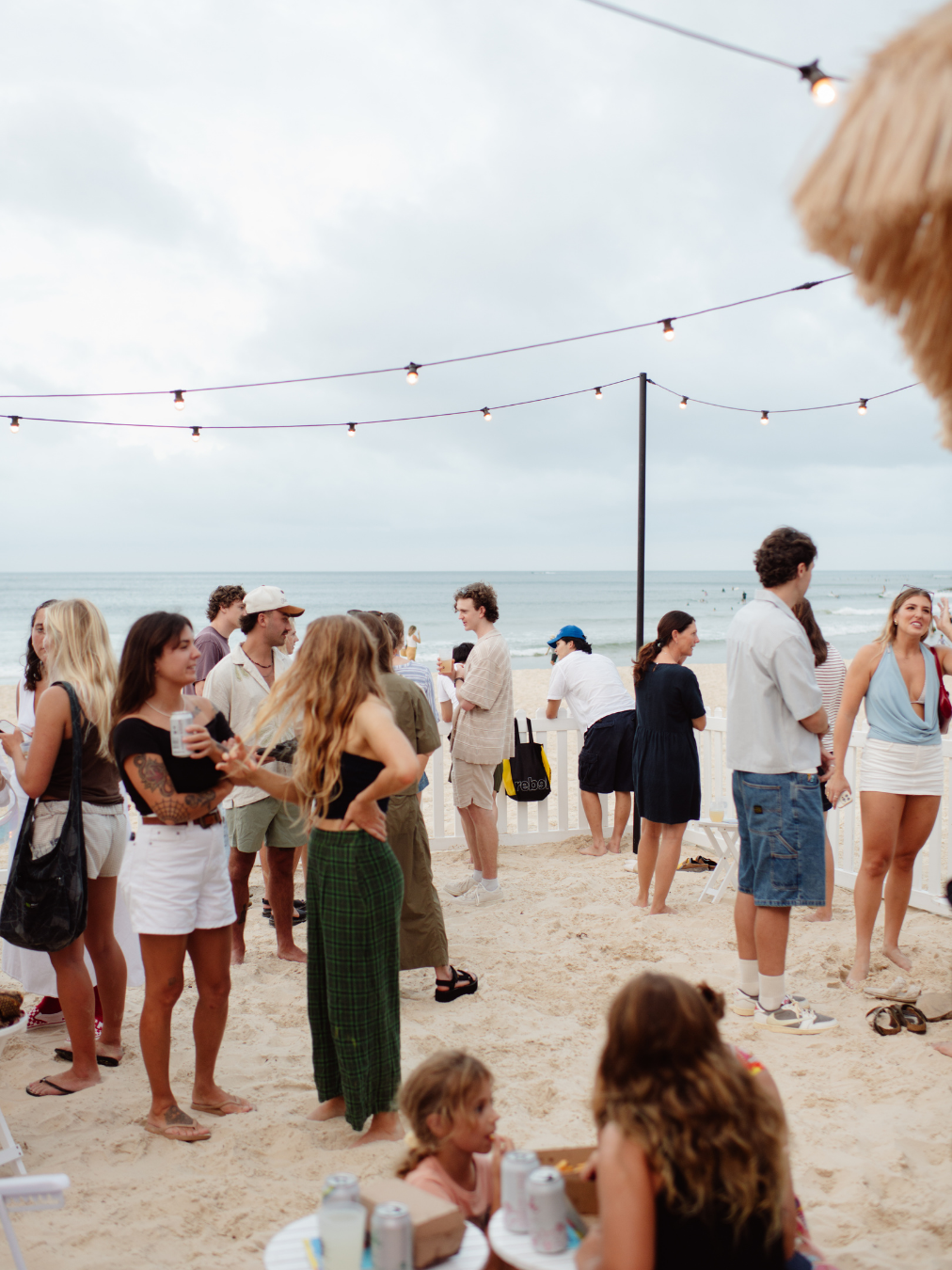 Group of people gathered on a beach under string lights, with the ocean in the background, some holding drinks and socializing.