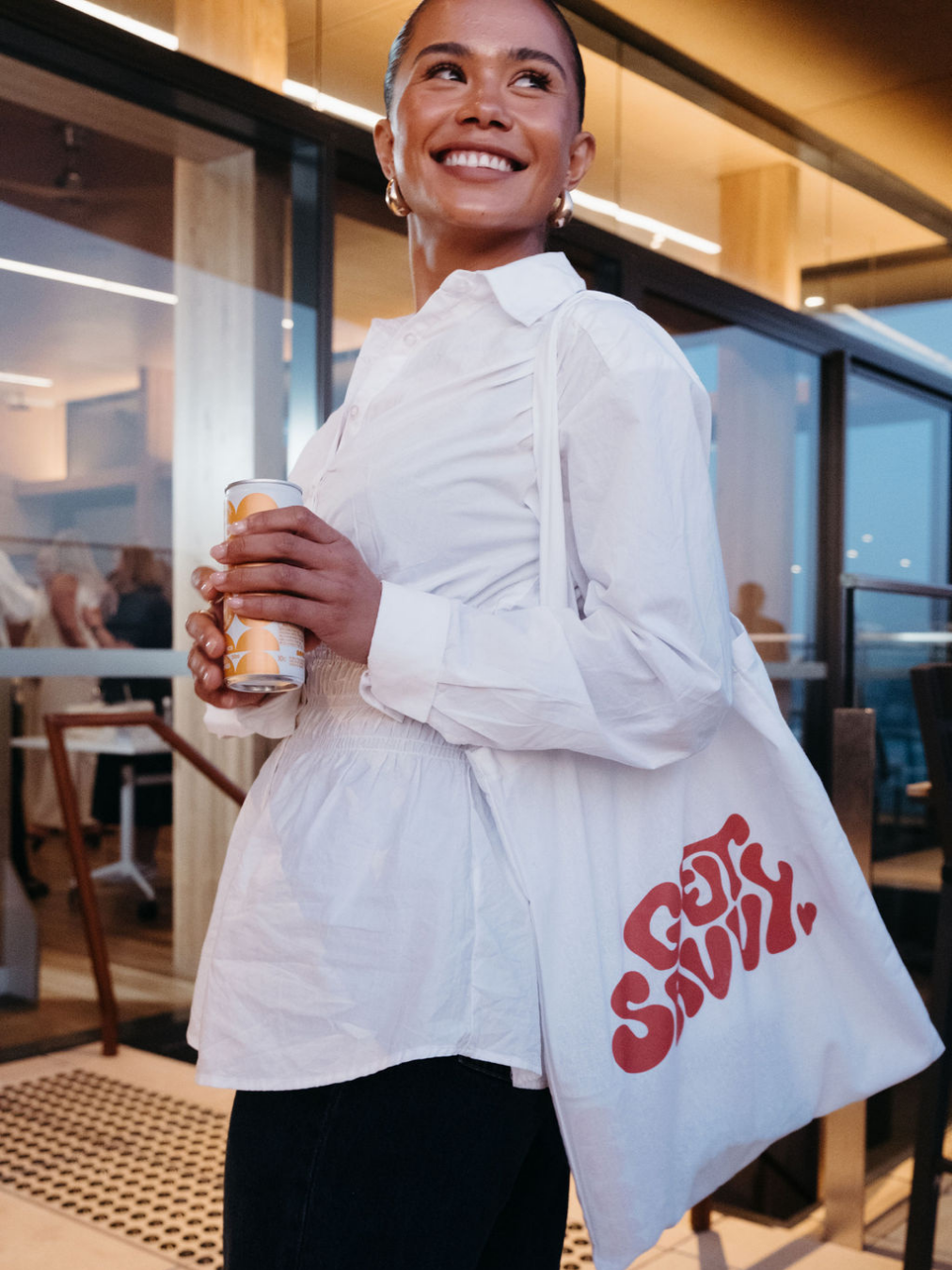 A woman smiling and holding a can of beverage, standing indoors with a large bag that has 'SASSY SASSY' printed on it.