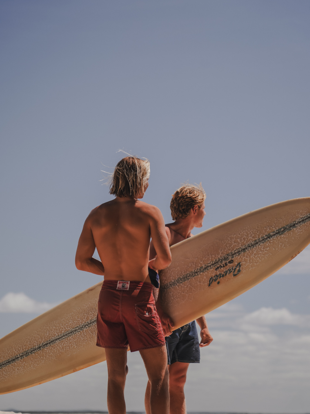 Two shirtless men, one with long blonde hair and the other with short blonde hair, holding a surfboard on a beach, with a clear blue sky in the background.