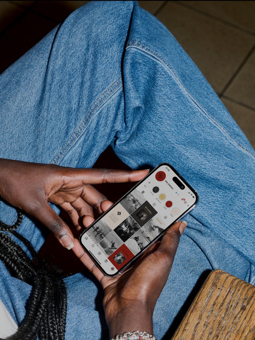A person with dark skin wearing blue jeans, holding a smartphone with a music app open, sitting on a wooden surface.
