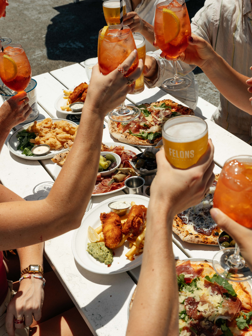 People enjoying a meal outdoors with drinks, pizza, fried chicken, and various appetizers.