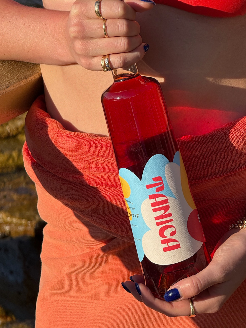 Close-up of a woman holding a bottle of cherry-flavored sparkling water outdoors. She is wearing rings and has dark blue nail polish. The woman is dressed in a red-orange top, and the background includes a rocky, natural setting with sunlight.