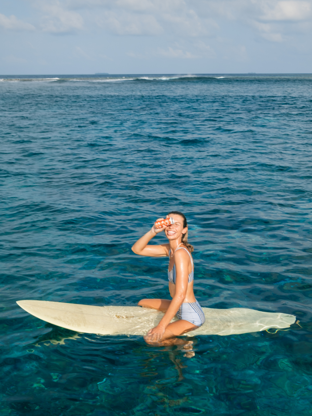 A woman sitting on a surfboard in the ocean, smiling and holding a drink, with clear blue water and a partly cloudy sky.