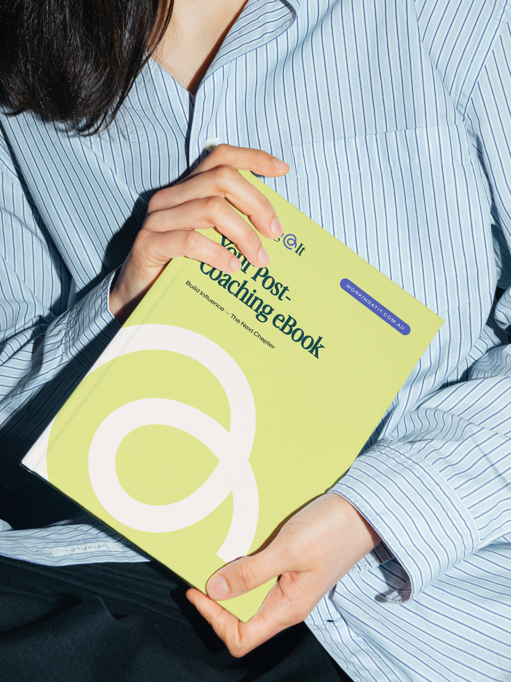 Person wearing a blue and white striped shirt holding a green book titled 'Your Post-Coaching eBook' standing.