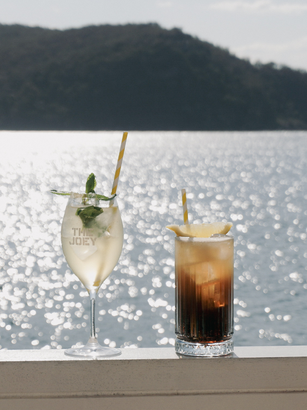 Two cocktails on a railing by the water with an island in the background, one with lime, mint, and a striped straw, and the other with lemon and ice, both glistening in sunlight.
