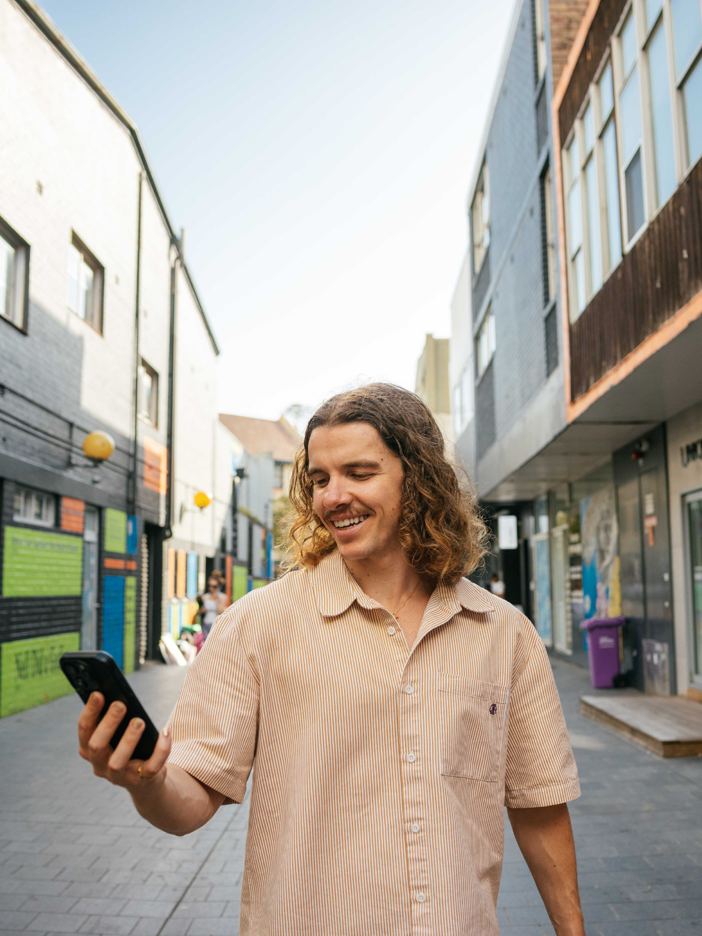 A young man with curly hair smiling and looking at his phone while walking through a city street lined with modern buildings and colorful wall art.