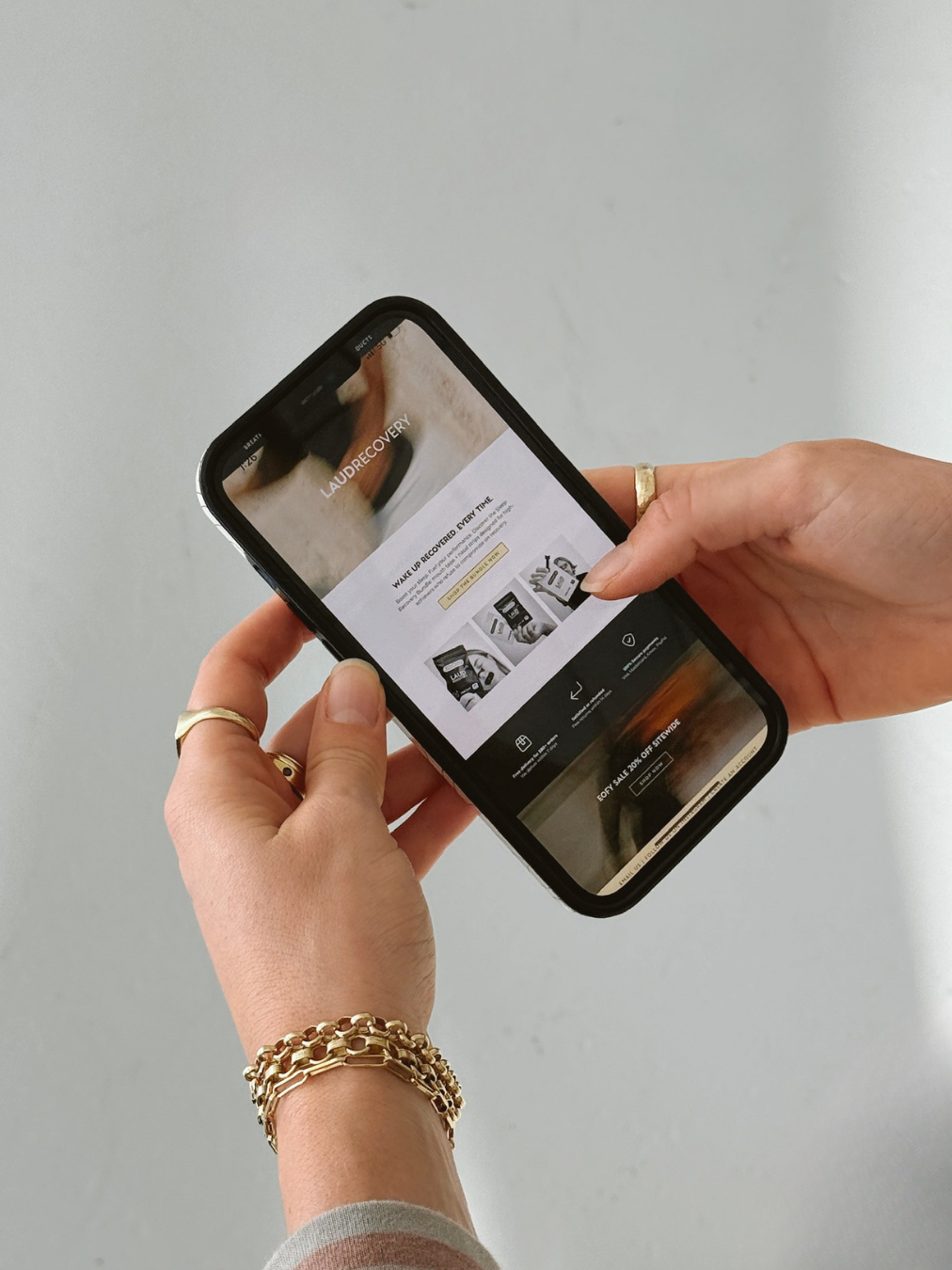 Person holding a smartphone viewing the Laudrecovery website with jewelry on their hands, wearing multiple gold bracelets and rings, against a plain light background.