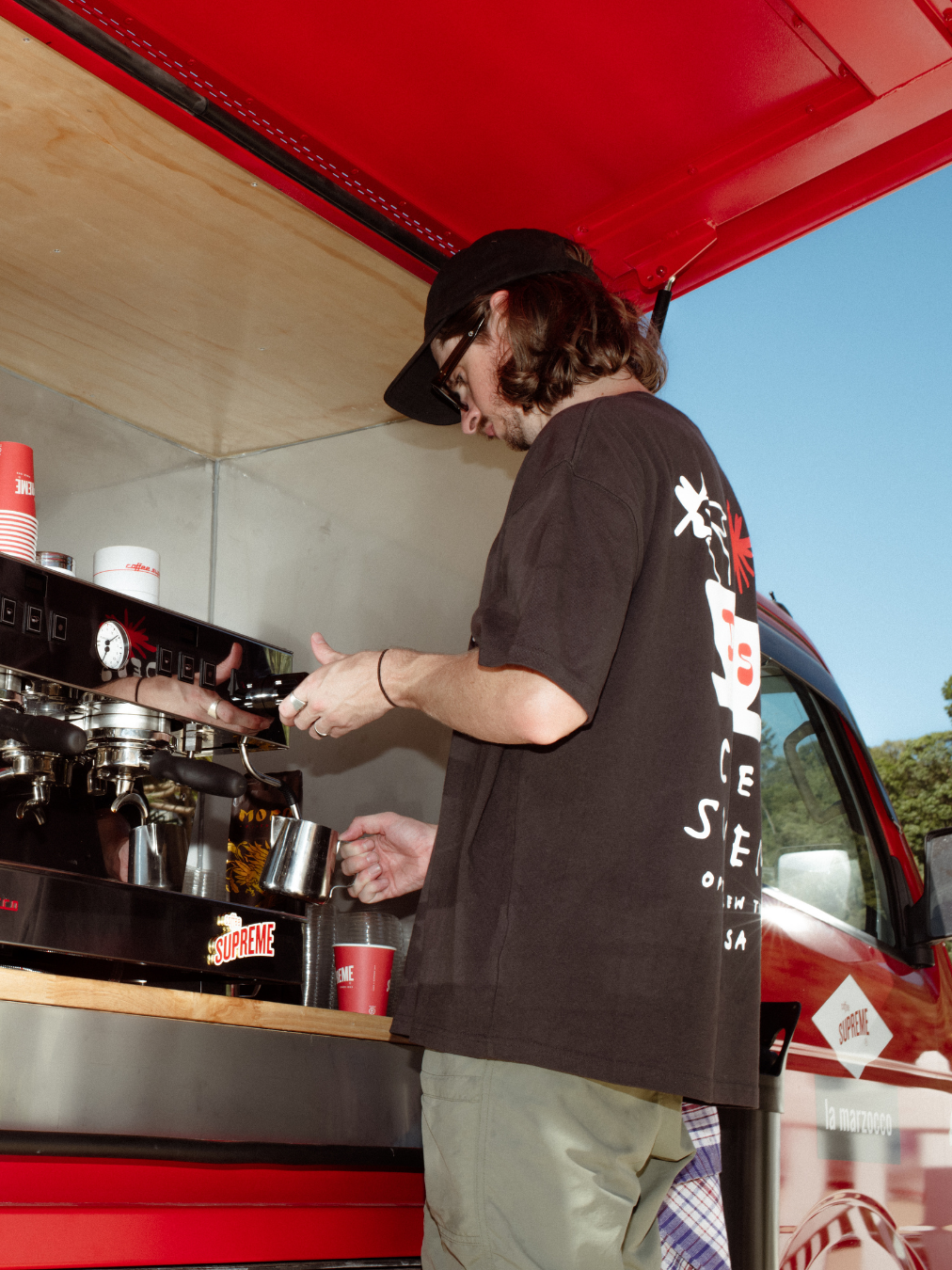 A person with long hair, black cap, sunglasses, black t-shirt, and beige pants making coffee with an espresso machine inside a red food truck.