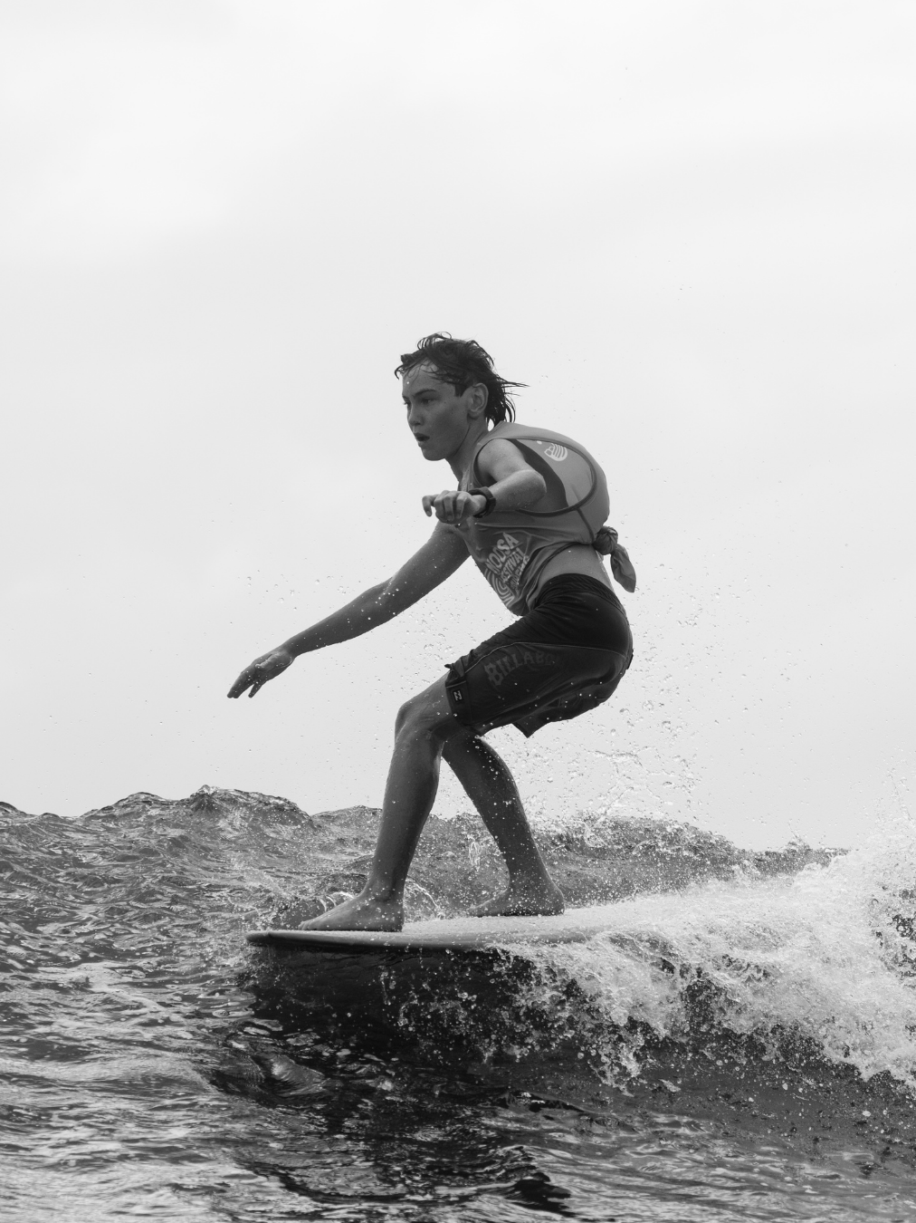 A person surfing on a wave, wearing a life vest and shorts, with wet hair, captured mid-ride on a black and white photo.