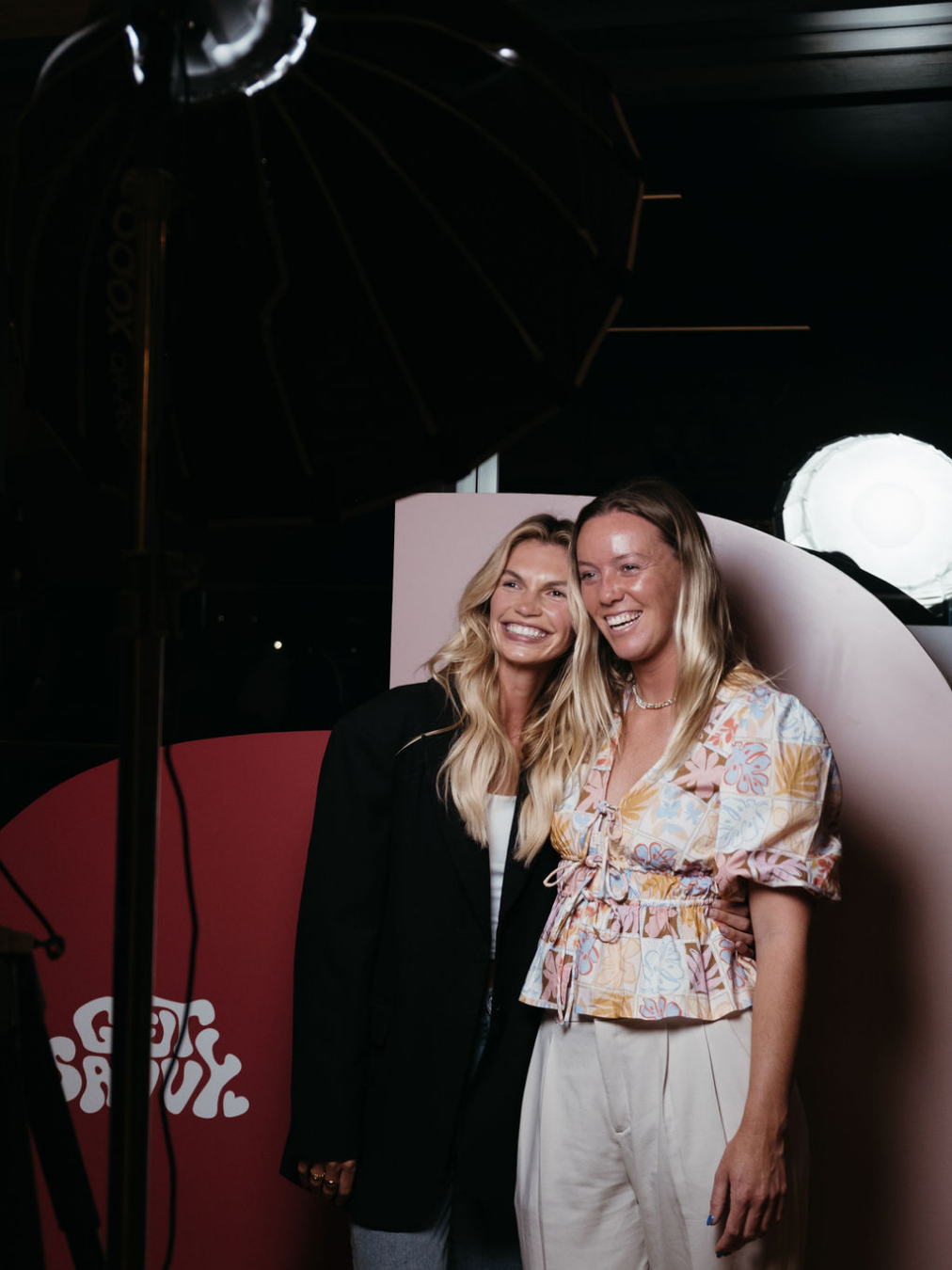 Two women smiling and posing together in front of a photo backdrop with studio lighting and a large umbrella reflector.