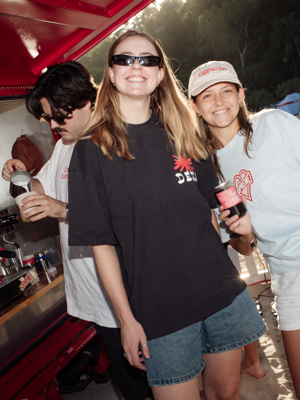 Three young women smiling at an outdoor event, one holding a cup and another holding a small jar, with a man making coffee in the background, surrounded by trees and tents.
