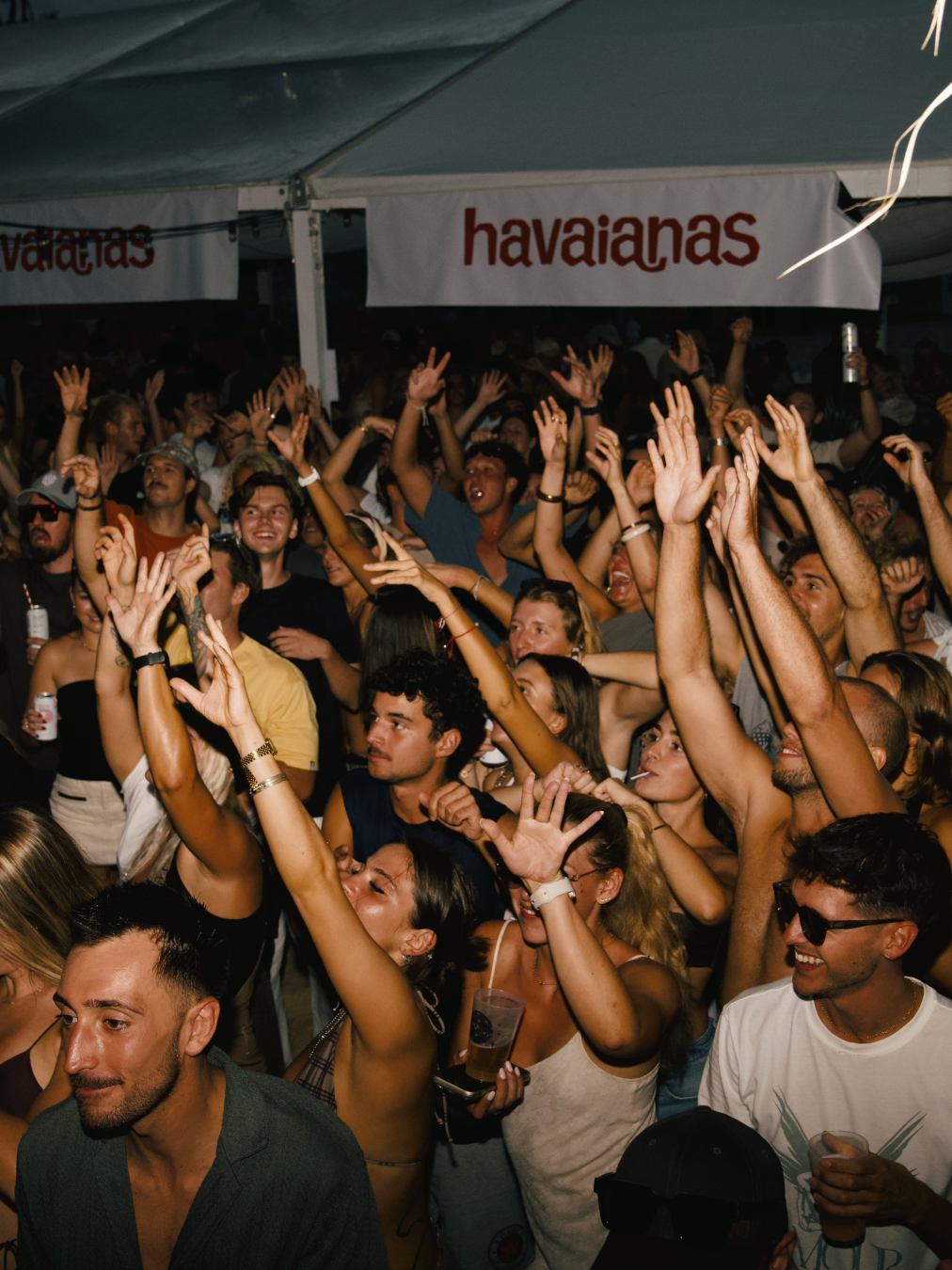 Crowd of people dancing and having fun at a party or concert inside a tent, with a banner that reads 'havaianas' in the background.