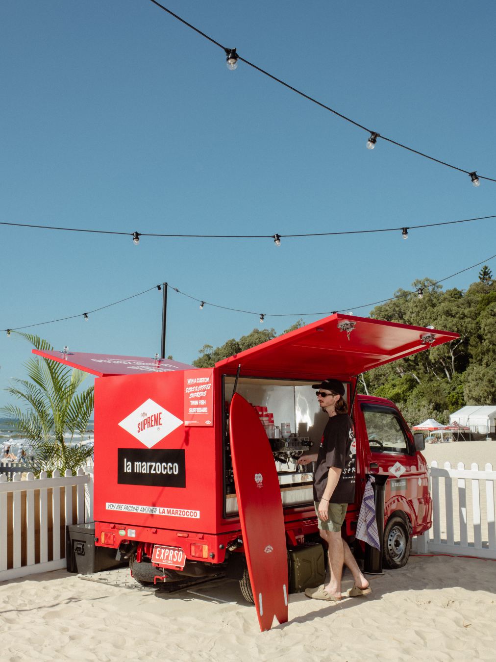 A red food truck named 'la marzocco' serving coffee at a beach with string lights overhead and palm trees in the background.