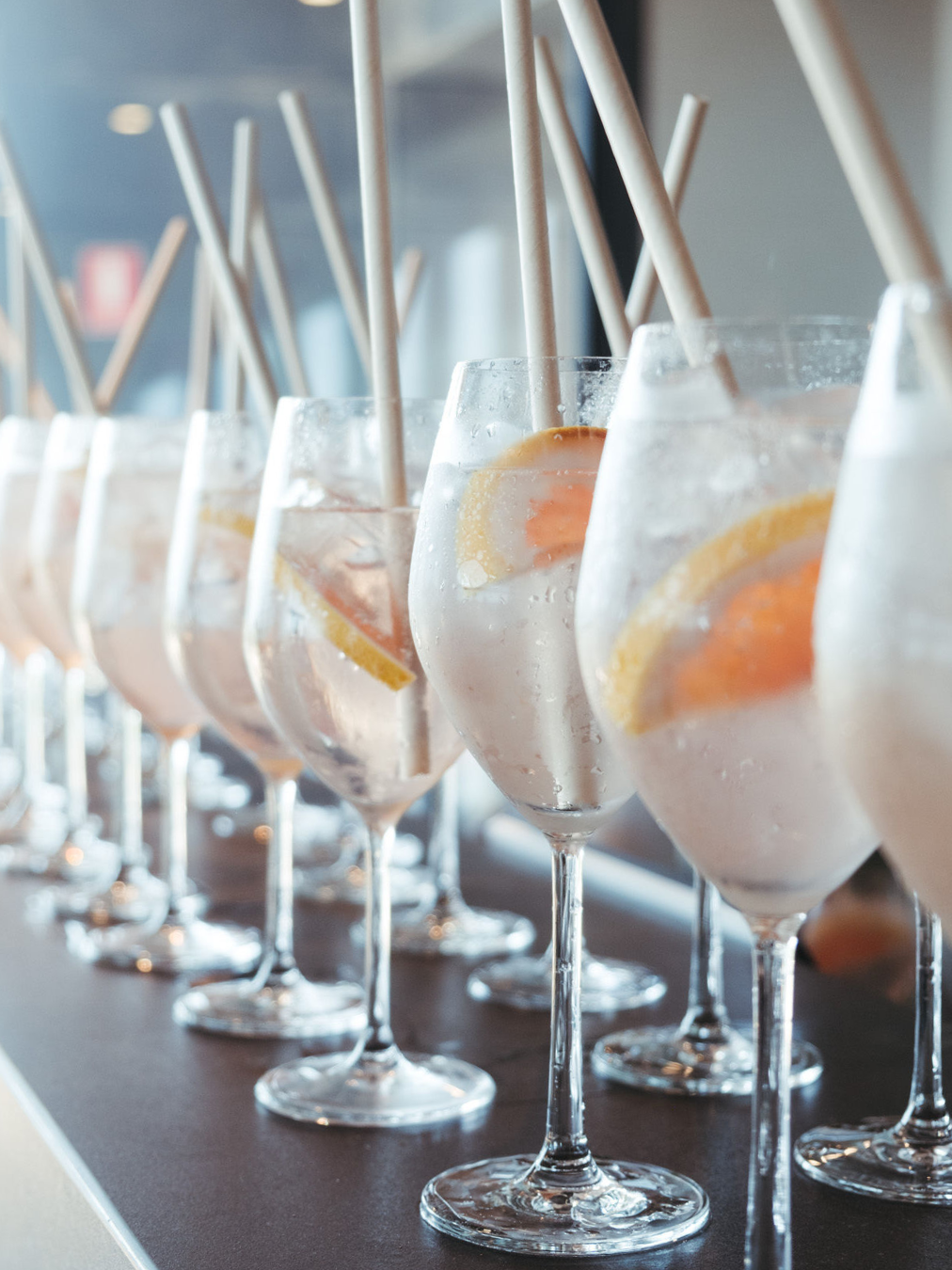 A row of tall glasses filled with a clear beverage, ice, and lemon slices, with straws, on a bar or counter.