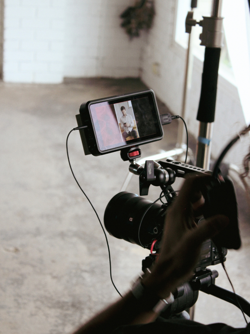 A videographer operating a camera on a tripod, with a monitor displaying a person sitting against a white brick wall.