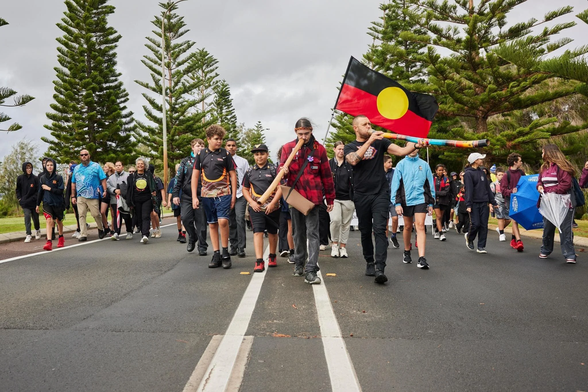 A diverse group of people, including children and adults, walking down a street lined with tall pine trees. One man is playing a didgeridoo, and another person is holding an Aboriginal flag. The scene suggests a cultural or community event.