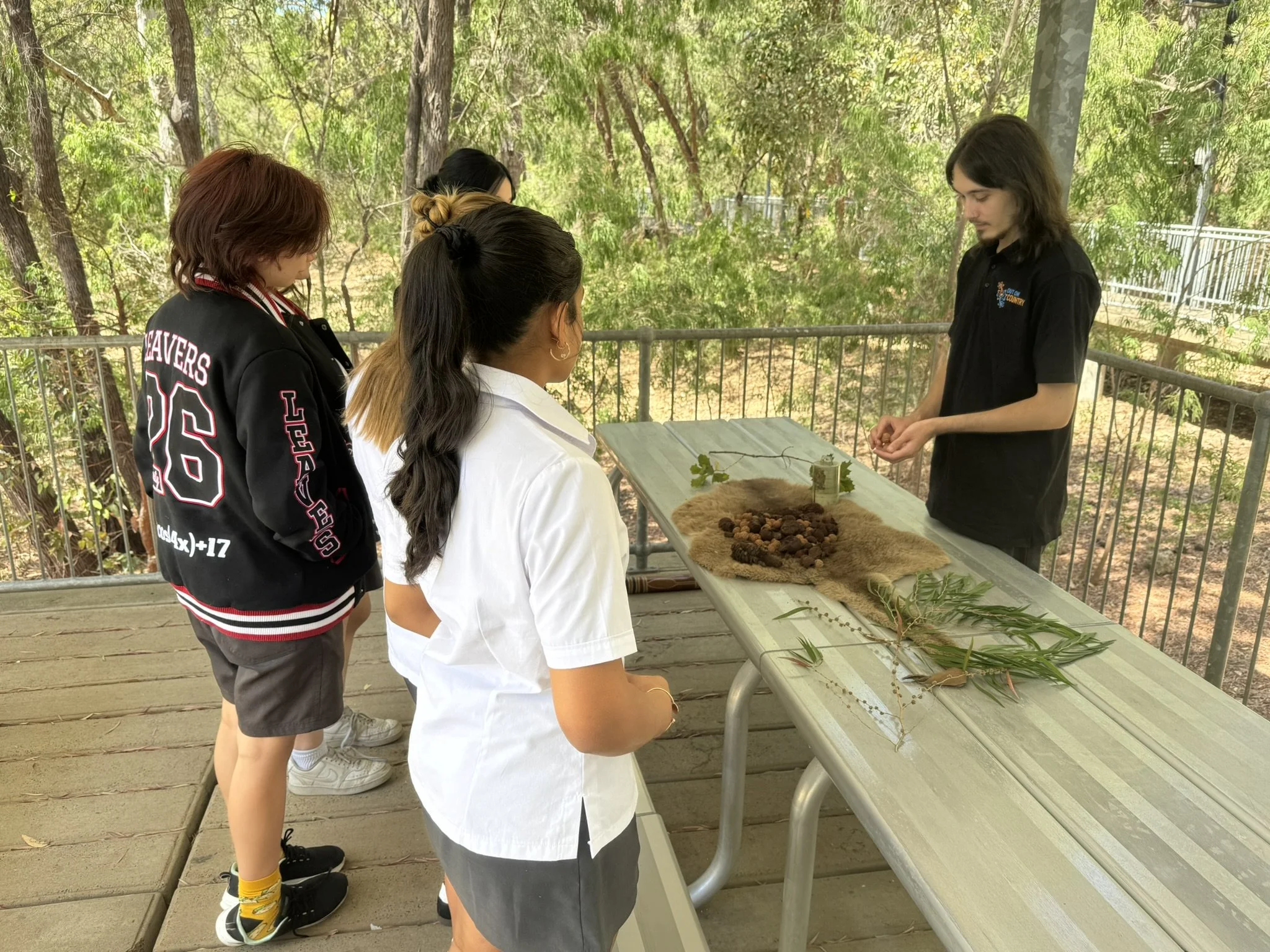 Group of children observing objects on a table outdoors, possibly a nature or educational activity, with trees and greenery in the background.
