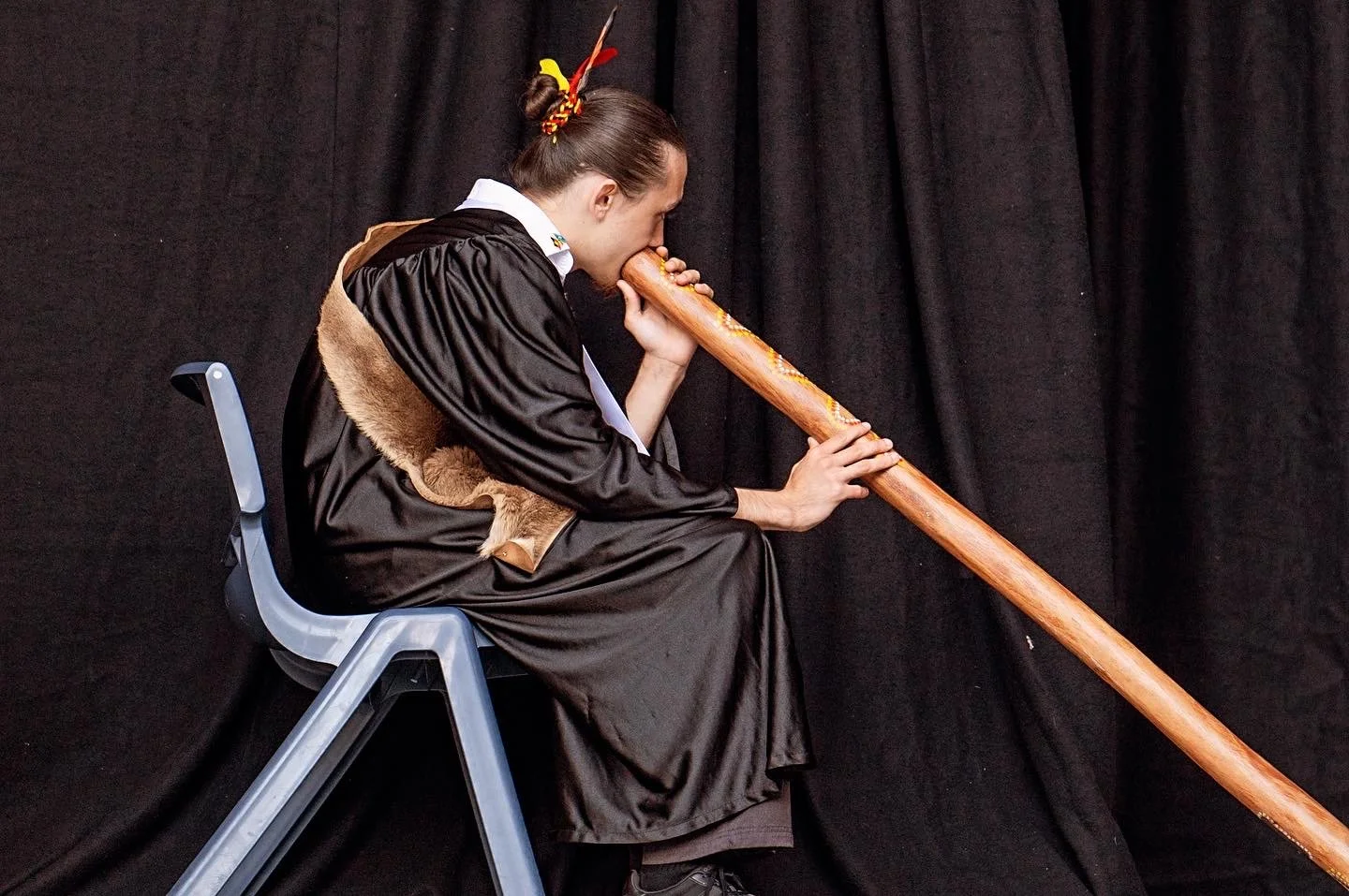 A woman dressed in academic robes plays a didgeridoo while sitting on a chair against a black curtain background.