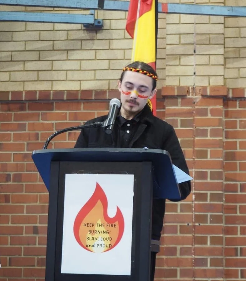 A young person with face paint and a braided headband speaks at a podium with a fire flame graphic, inside a room with brick walls and a flag in the background.