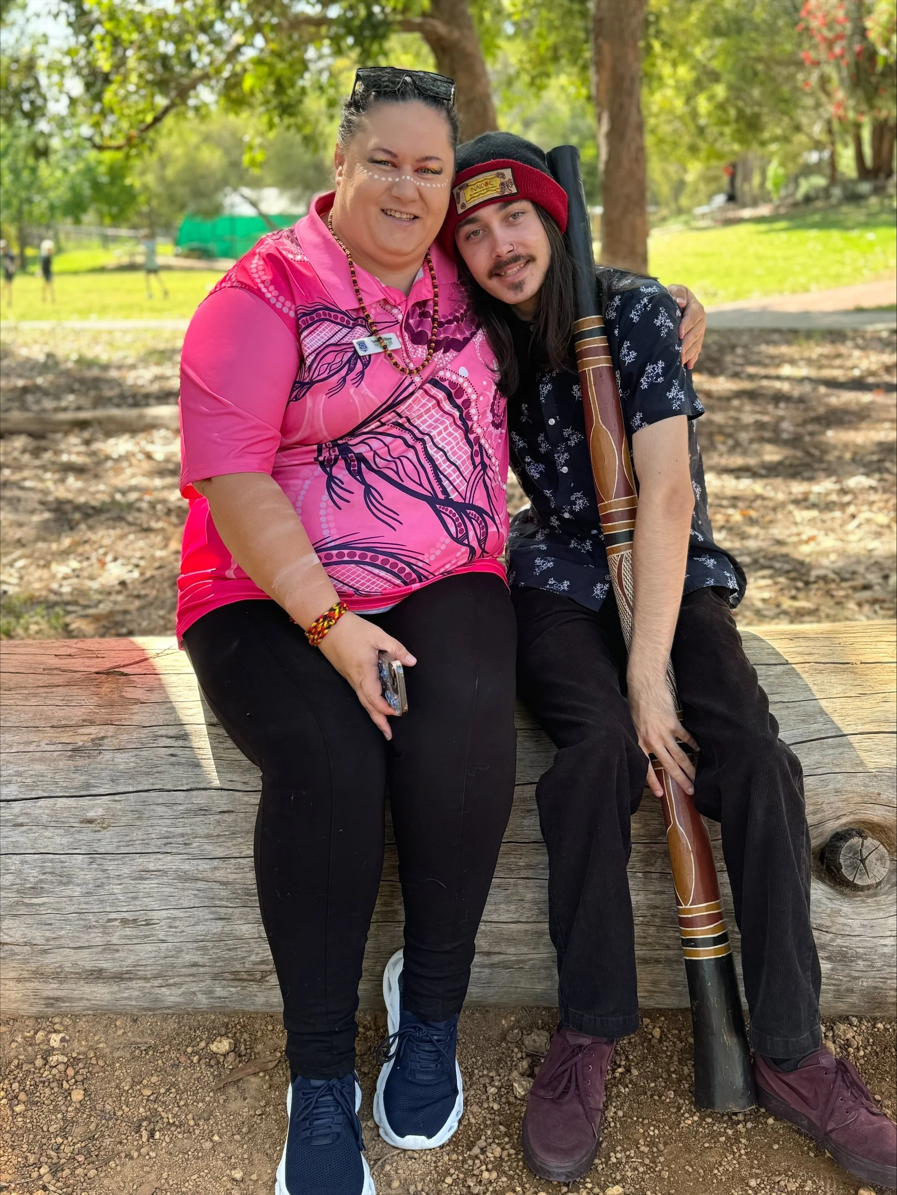 Two people sitting closely together outdoors on a wooden bench, one person holding a wooden flute. They are smiling, with a background of trees and greenery.