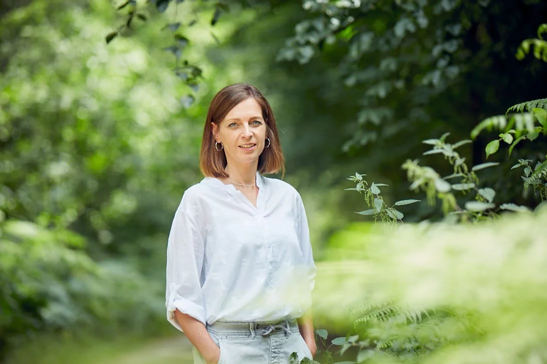 Frau steht in einem grünen, bewaldeten Außenbereich, trägt weiße Bluse und Jeans, lächelt freundlich.