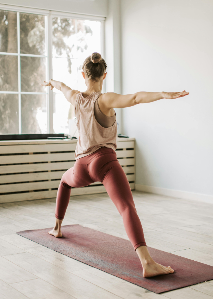 Frau macht Yoga im Wohnzimmer auf Yogamatte, schaut auf die Fensterfront.