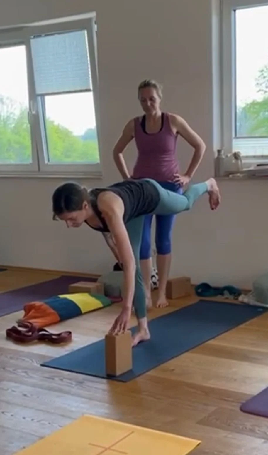 Zwei Frauen üben Yoga in einem Raum mit großen Fenstern, die natürliches Licht hereinlassen. Eine Frau steht hinter der anderen, die sich in eine Balancierübung im Handstand befindet, wobei sie die Balance auf Holzbalken hält.