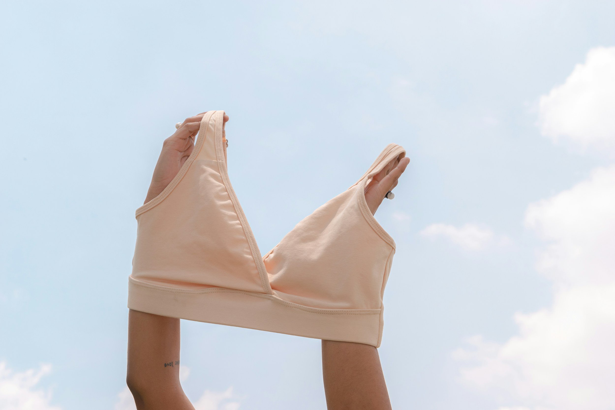 A person holding a beige sports bra against a sky background with clouds.