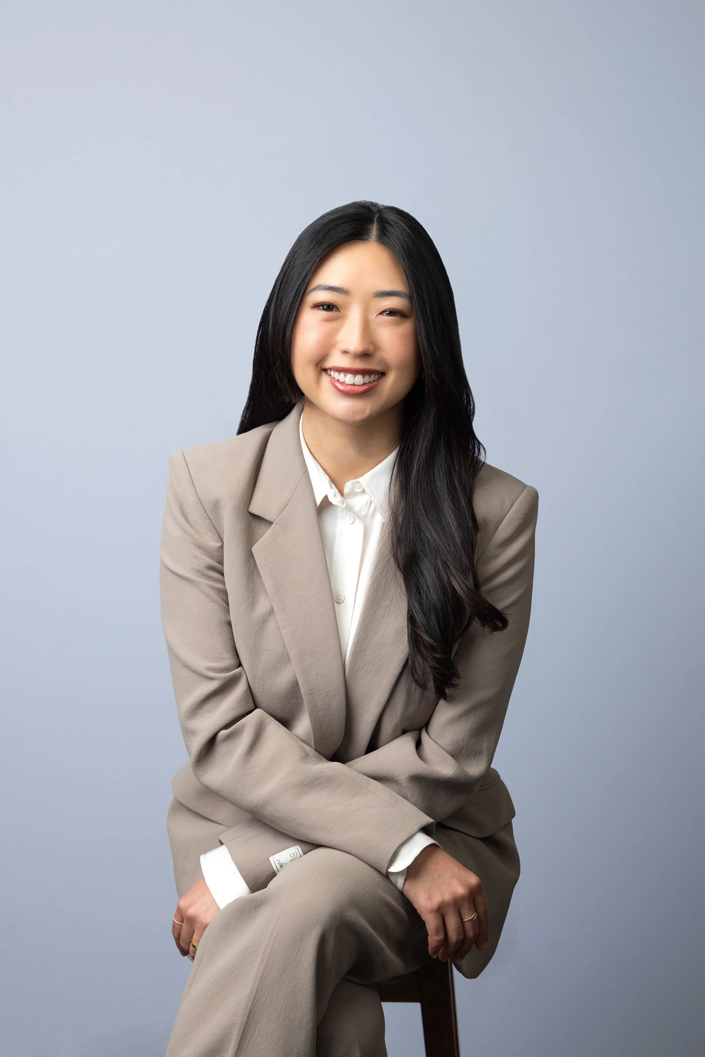 A woman with long black hair, wearing a beige suit and white shirt, sitting on a wooden chair, smiling with one eye winking against a plain light gray background.
