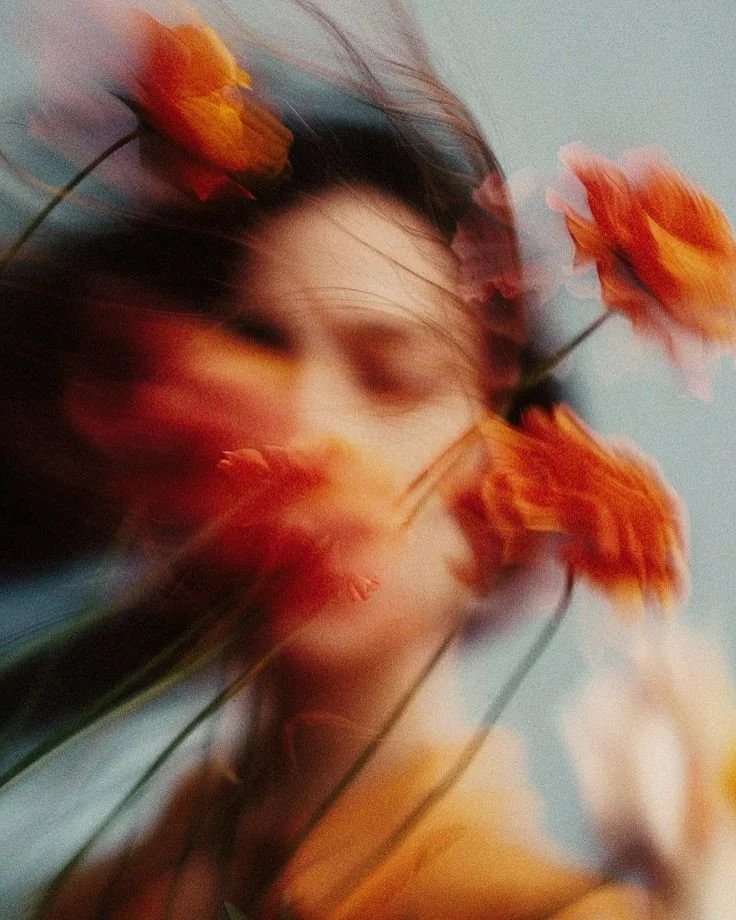 Blurred photo of a person with long hair and several orange flowers covering their face, against a sky background with clouds.