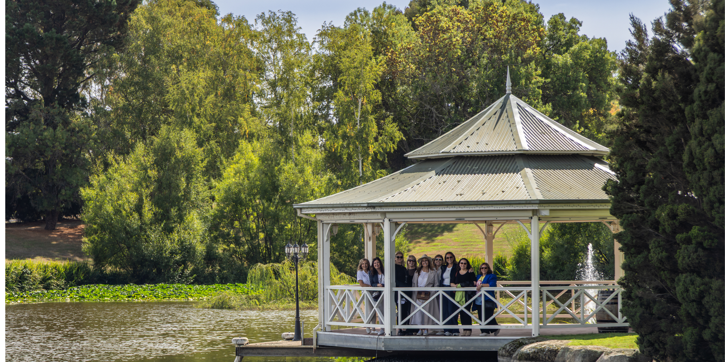 Group of people standing on a pavilion near a pond surrounded by green trees.