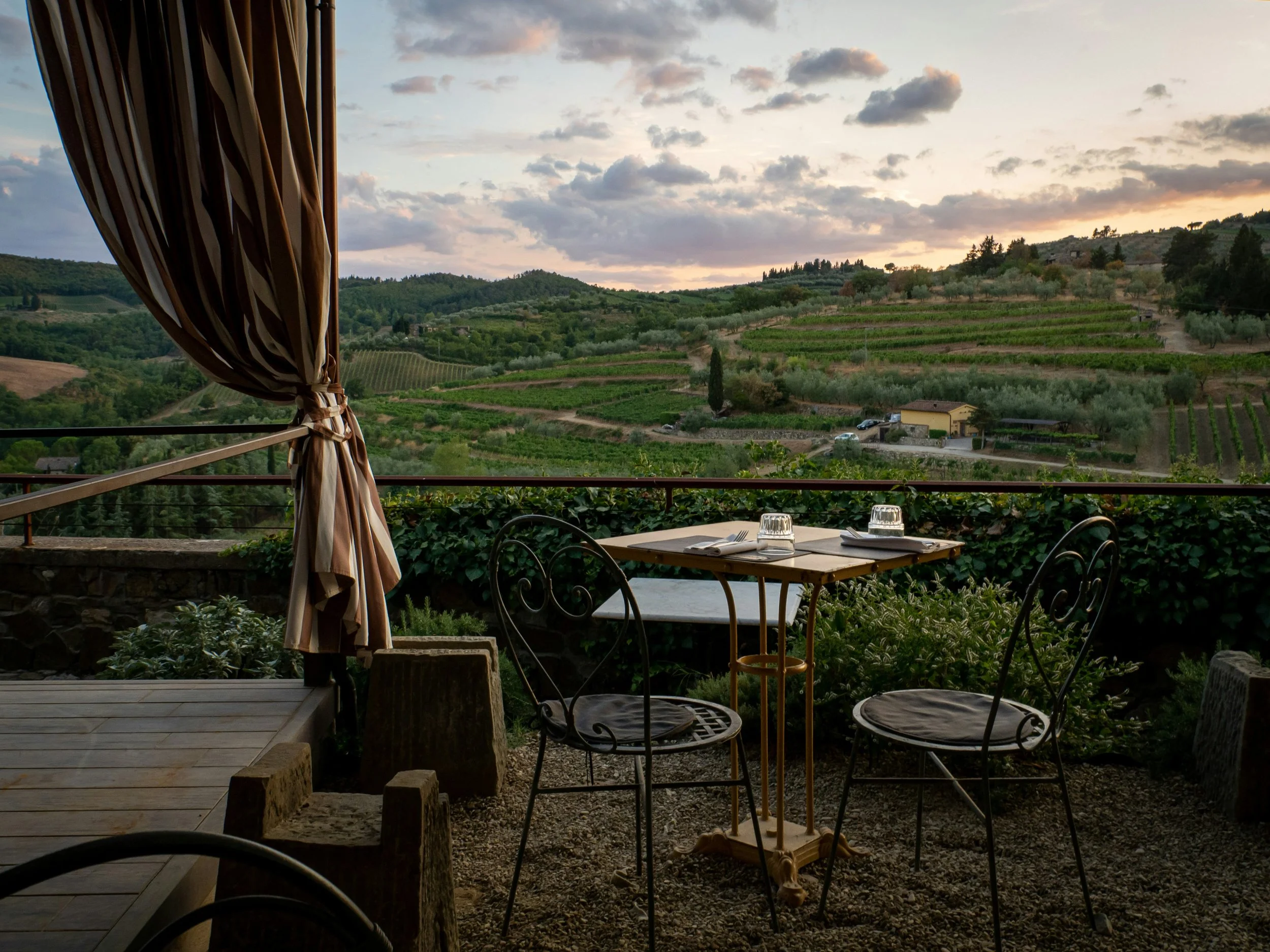 View from a restaurant or cafe terrace overlooking rolling green hills with vineyards, trees, and a few small buildings at sunset, partially shaded by a striped curtain.
