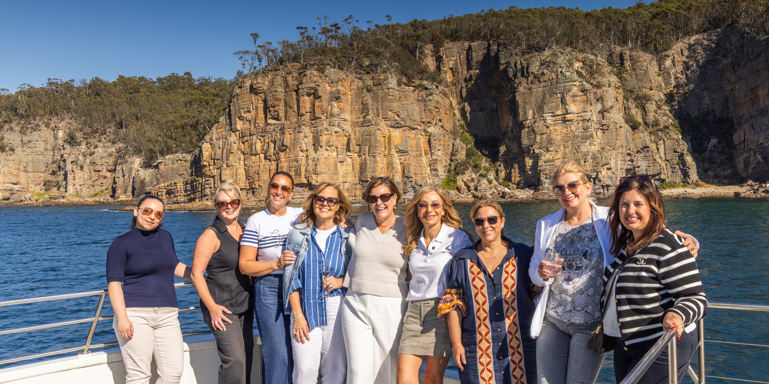 A group of women standing on a boat with a rocky cliff and trees in the background, enjoying a sunny day on the water.