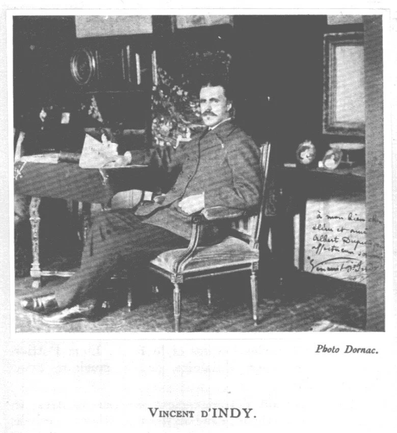 A black and white photograph of a man sitting on a chair in a room, with various objects and framed pictures in the background, and a handwritten note on the right side of the image.