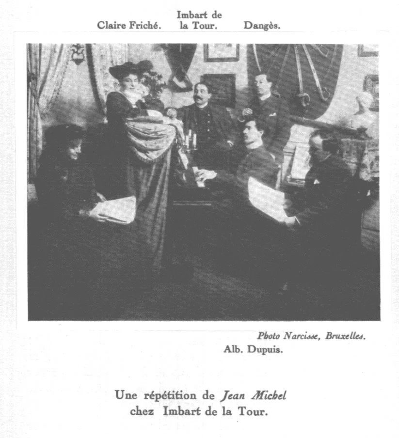 A black-and-white photograph of six people gathered around a table in a room with paintings and a large clock on the wall. The scene appears to be a theatrical or artistic setting. Text above the image reads 'Claire Friché. Imbart de la Tour. Danges.' and below the photo notes 'Photo Narcisse, Bruxelles. Alb. Dupuis.' with a caption in French indicating a rehearsal of Jean Michel at Imbart de la Tour.