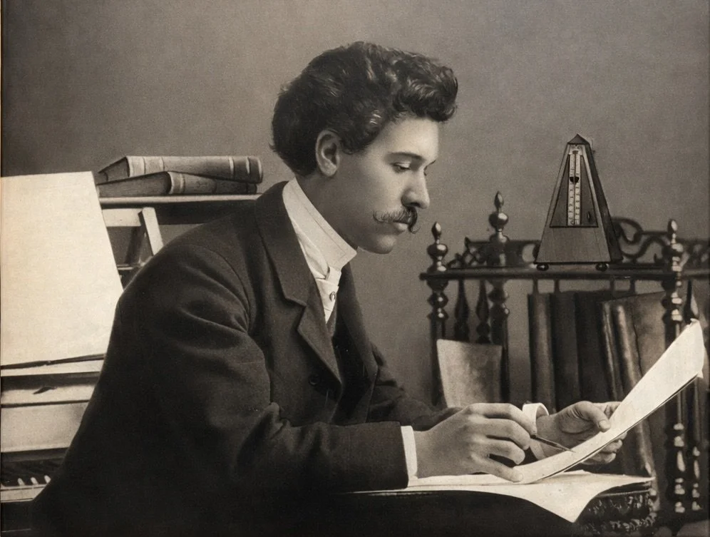 A black-and-white portrait of a young man with dark, wavy hair and a mustache, reading a book or document, seated at a desk with books and a metronome in the background.