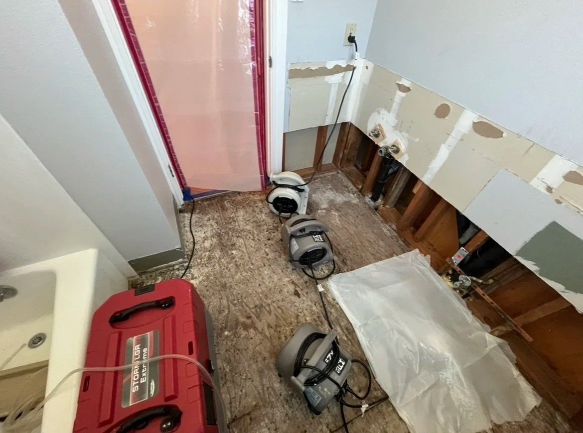 A room under renovation with exposed flooring and wall pipes, with four wet/dry shop vacuums and a red portable generator on the floor, and a plastic sheet covering part of the floor.
