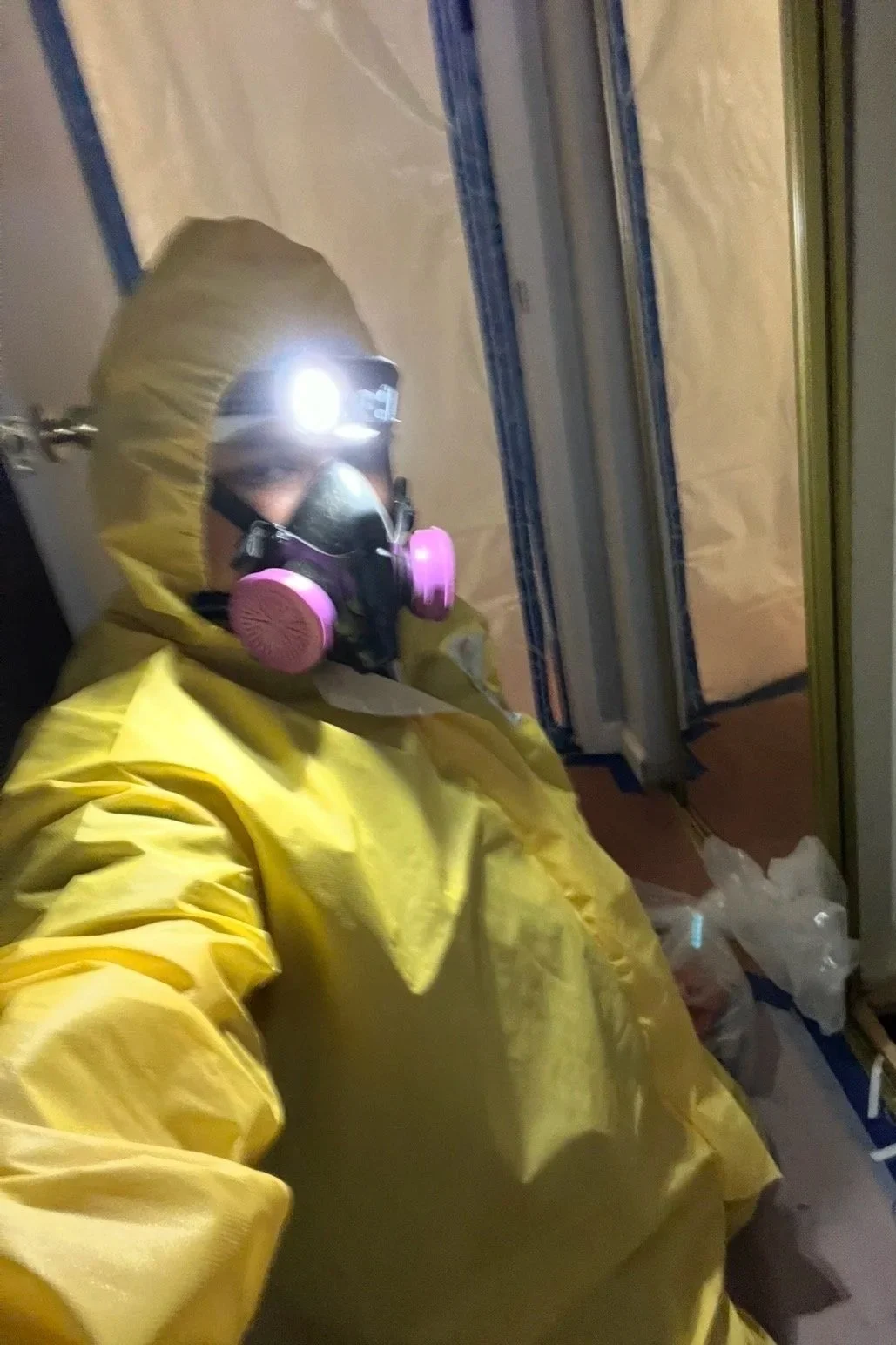 Person wearing yellow protective suit, pink respirator mask, and headlamp, in a construction or sanitation setting.
