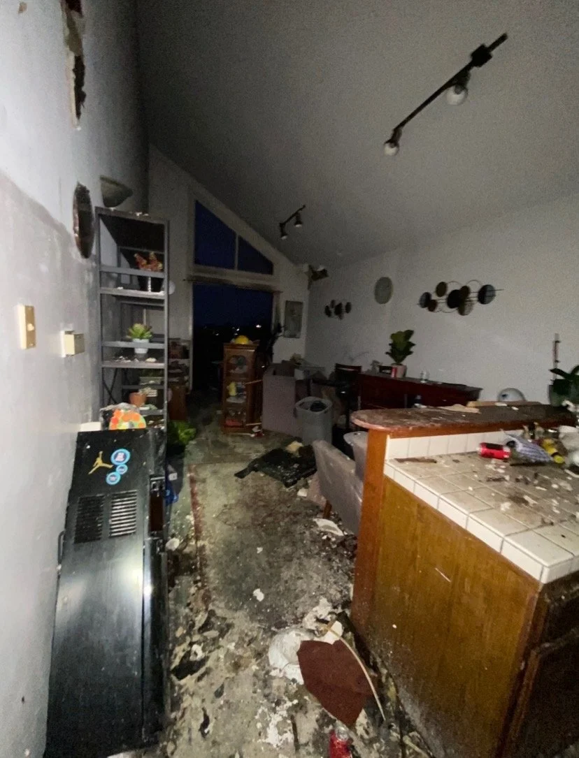 Living room and kitchen area damaged by fire with debris, soot, and burnt furniture and belongings scattered around.