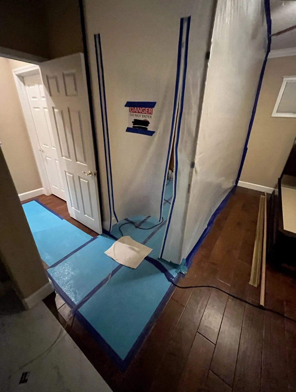 Room undergoing renovation with contaiment, taped at the edges, and a caution sign, with a small cloth on blue protective flooring.