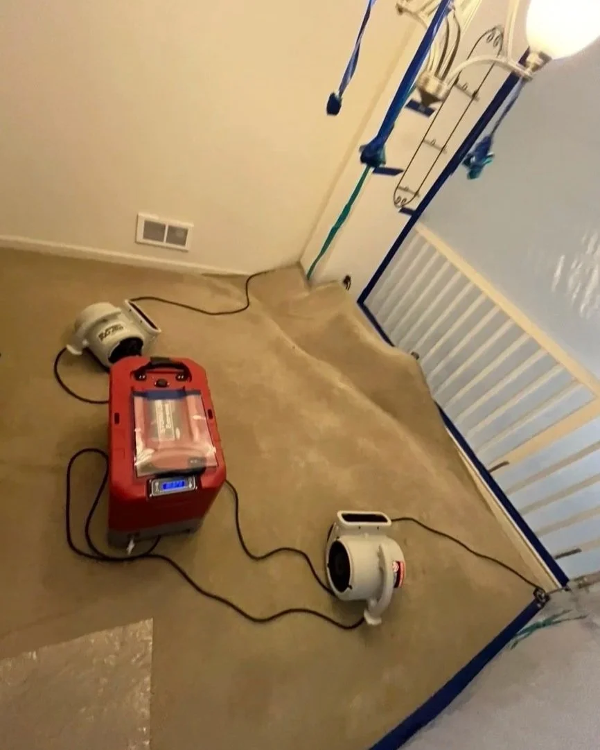 A room with a painted floor, a white and red device connected to two white air movers, plastic sheeting, and painter's tape on the walls, indicating ongoing painting or renovation work.