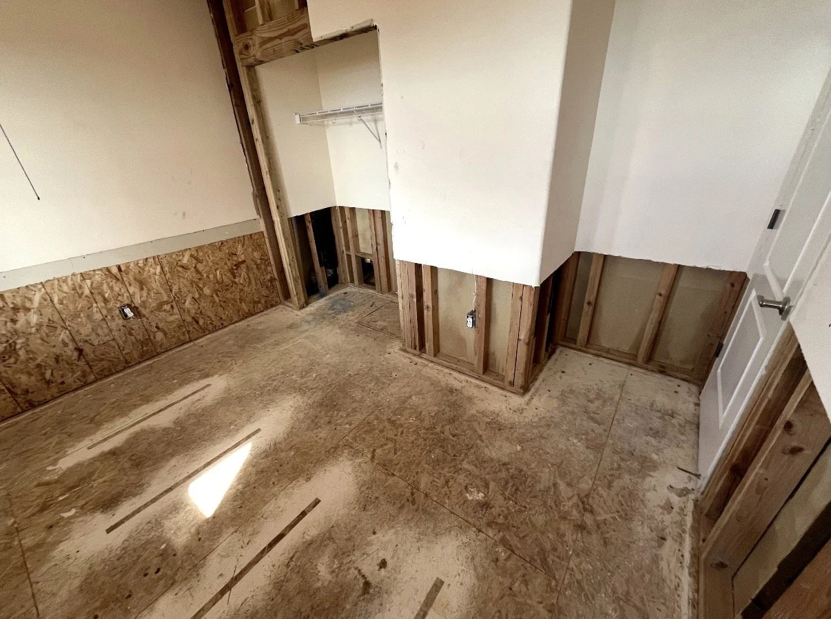 An unfinished room with exposed wooden framing and OSB plywood on the floor. The walls have unfinished drywall, with some electrical outlets visible. There is a small closet space with a wire shelf, and a white door on the right side.