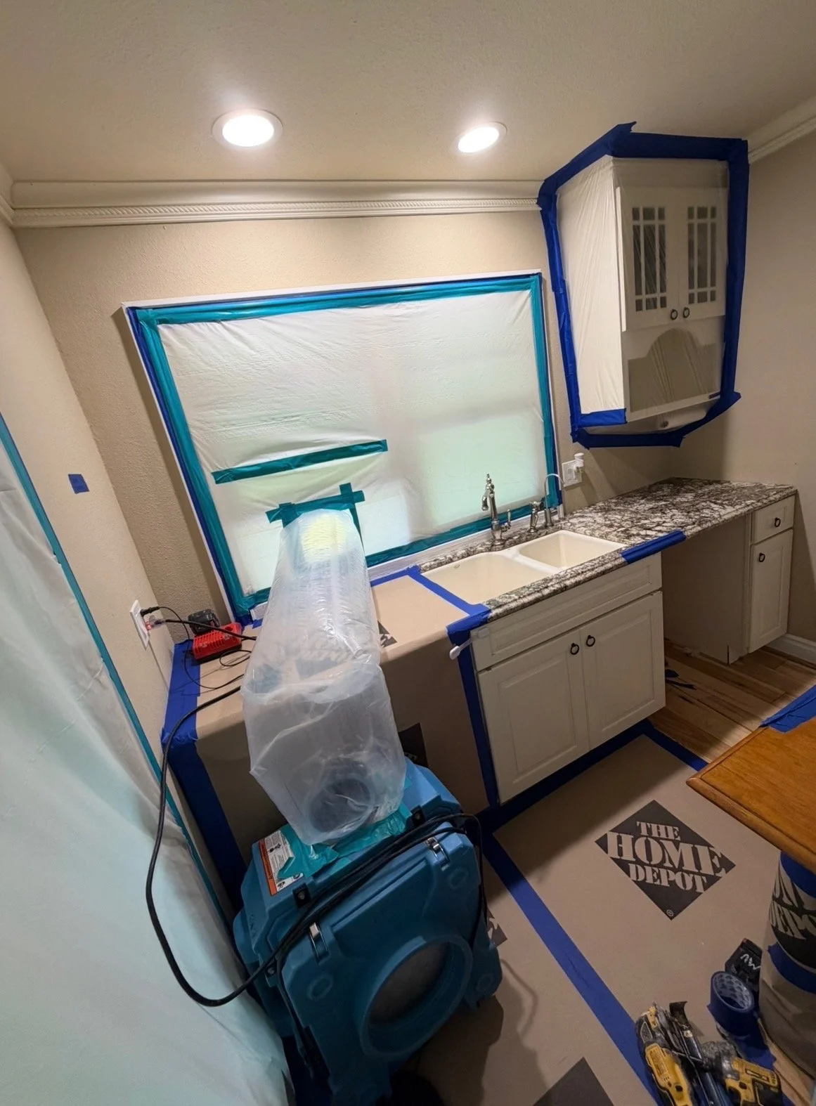 Kitchen with cabinets, granite countertops, double sink, and window covered with plastic and blue painter's tape for painting or renovation work, with tools and equipment nearby.