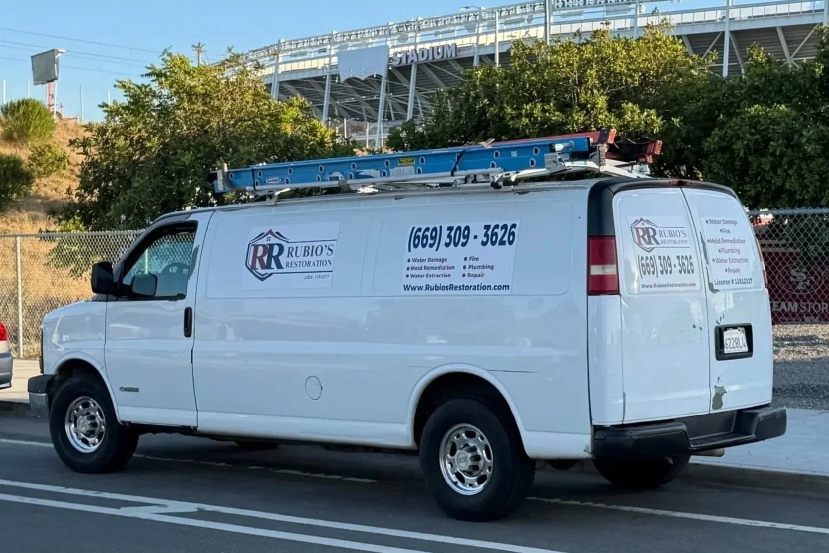 White Rubio's Restoration service van with blue and black lettering parked near a chain-link fence, with trees and a stadium in the background.