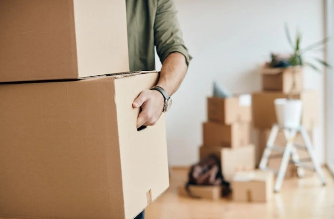 Person holding a large cardboard moving box in a room with more boxes and a small ladder in the background.