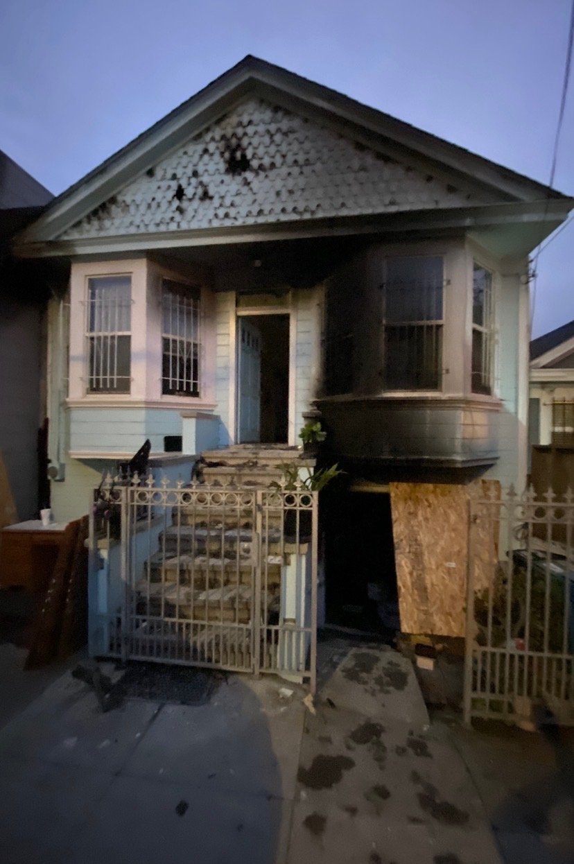 Damaged house with boarded-up front entrance and burned sections, including charred windows and roof, with a metal gate at the front.